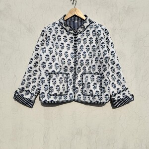 May include: A quilted jacket featuring a white base with a repeating dark blue floral block print pattern. The jacket has a mandarin collar, long sleeves with striped cuffs, a front button closure, and two square patch pockets with striped trim.