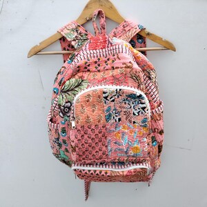 May include: A pink patchwork backpack with a white zip and straps, hanging on a wooden hanger. The backpack features various floral and geometric patterns in shades of pink, green, and blue. The bag has a front pocket and side pockets.