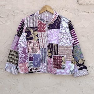 May include: A quilted jacket in a patchwork design, featuring various shades of purple, lavender, and white. The jacket has a collared neckline and is hung on a wooden hanger. The fabric includes floral and geometric patterns.