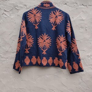 May include: A navy blue jacket with intricate coral-colored floral embroidery. The jacket features a mandarin collar and long sleeves, with a repeating floral pattern across the back and sleeves. The bottom hem is adorned with a row of coral-colored pomegranate-like designs.