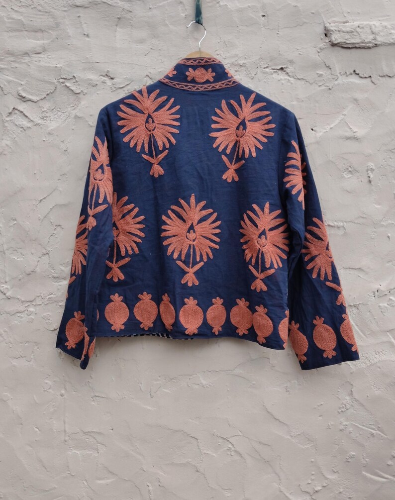 May include: A navy blue jacket with intricate coral-colored floral embroidery. The jacket features a mandarin collar and long sleeves, with a repeating floral pattern across the back and sleeves. The bottom hem is adorned with a row of coral-colored pomegranate-like designs.