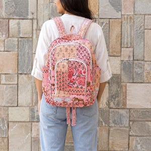 May include: A quilted backpack featuring a patchwork design in shades of pink, peach, and cream with floral and botanical patterns. The bag includes a front zippered pocket, two side pockets, and adjustable shoulder straps with white stitched edges.