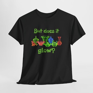 May include: A black short-sleeve t-shirt featuring a graphic of various colorful glass figurines and bowls. The text "But does it glow?" is printed in lime green above and below the collection of glass items in the center of the shirt.