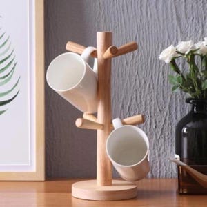 Puede incluir: Un árbol de tazas de madera con una base redonda y múltiples clavijas para colgar tazas. Dos tazas de cerámica blanca se exhiben en el árbol. El árbol está hecho de madera clara.