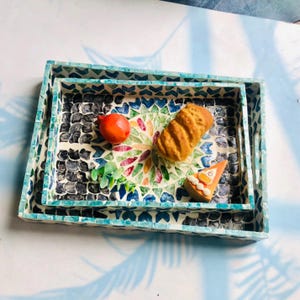 May include: Nested decorative trays with mosaic tile design. The trays have a turquoise and white border, and the interior features a colorful floral pattern. The trays contain a red fruit, a pastry, and a small plant.