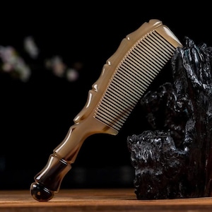 Puede incluir: Un peine artesanal con un acabado pulido de color marrón claro. El peine presenta un mango decorativo con un diseño curvo y dientes finos. El peine está hecho de un material natural, posiblemente cuerno o madera, y se muestra sobre un fondo oscuro.