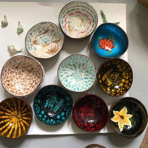 Puede incluir: Una colección de cuencos decorativos de coco con varios diseños. Algunos cuencos presentan patrones de mosaico, mientras que otros muestran imágenes pintadas de tortugas, flores y otros motivos tropicales. Los cuencos son de varios colores, incluyendo azul, verde y amarillo.