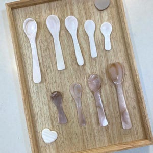 May include: A collection of decorative spoons in various sizes and colors, arranged on a wooden tray. The spoons range from white to light brown, with a pearlescent sheen. Two heart-shaped decorative pieces are also present.
