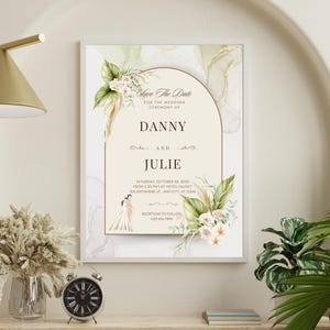 May include: A framed wedding invitation with the text "Save The Date" and the names Danny and Julie. The invitation features floral and leaf accents, with a neutral color palette. The invitation is displayed on a shelf with a clock and vase.