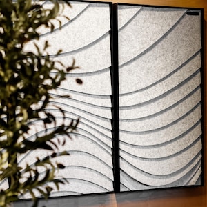 May include: Two framed wall panels with a wave-like design. The panels have a light gray, textured surface with curved, layered lines. The frames are black, and the panels are displayed against a light-colored wall.