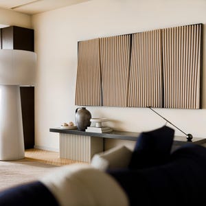 May include: A wall-mounted art piece consisting of four vertical panels with light wood slats, positioned above a long black console table. The table features a fluted base, a dark gray ceramic vase, a small white pedestal bowl, and a thin metal lamp.