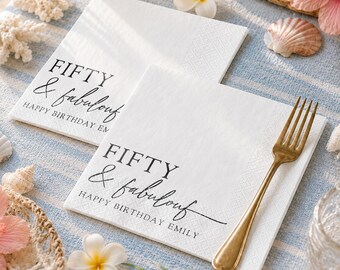 May include: Two square white paper napkins featuring black printed text that reads FIFTY & fabulous HAPPY BIRTHDAY EMILY. A gold-toned metal fork rests diagonally across the bottom napkin, which has a textured border and a smooth center.