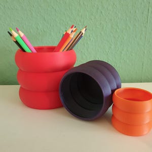 Könnte beinhalten: Ein roter, gestapelter, zylindrischer Stifthalter, gefüllt mit Buntstiften. Daneben befinden sich zwei weitere Halter: ein lila, gerippter Zylinder und ein orangefarbener, gestapelter, zylindrischer Halter. Die Halter stehen auf einer weißen Oberfläche mit grünem Hintergrund.