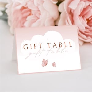 May include: A pink and white gift table sign with the words "GIFT TABLE" in brown lettering. Two pink butterflies are at the bottom of the sign. Pink flowers are in the background.