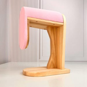 Puede incluir: Un expositor de joyas de madera con una parte superior acolchada de color rosa claro. El expositor está hecho de madera de color claro y tiene un diseño curvo. La base es rectangular y el expositor está diseñado para guardar pulseras o collares.