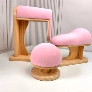 Könnte beinhalten: Drei Holz-Präsentationsständer mit rosa Samt-Oberflächen. Die Ständer haben unterschiedliche Formen: eine abgerundete Kuppel, eine geschwungene Form und eine rechteckige Form. Das Holz hat eine natürliche Oberfläche.