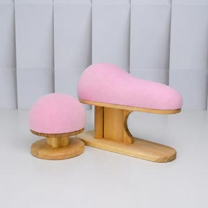 May include: A pink, mushroom-shaped hat block and a pink shoe last, both on wooden bases. The hat block is round, while the shoe last is elongated. The wooden bases have a natural finish. The background is a white, textured wall.