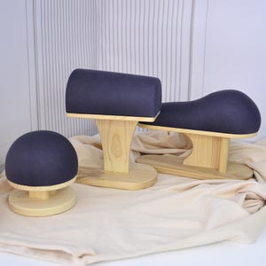 Op de afbeelding: Drie naaigereedschappen voor strijken met donkerpaarse, met stof beklede bovenkanten en lichtgekleurde houten voetstukken. De set bevat een rond kleermakerskussen, een cilindrische strijkrol en een gebogen mouwplank voor kleding.