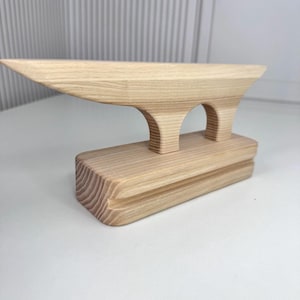 May include: A light-colored wooden ironing board with a rectangular base and a curved top. The board features two arched supports and a pointed end, showcasing the natural wood grain.