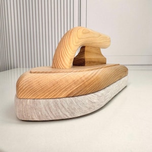 Könnte beinhalten: Ein hölzernes Bügelholz mit einer glatten Oberfläche aus hellem Naturholz. Das Werkzeug verfügt über einen geschwungenen, ergonomischen Griff, der auf einer massiven Basis mit flacher Unterseite befestigt ist, um Nähte zu glätten.