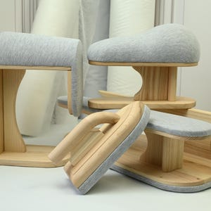 Op de afbeelding: Een verzameling houten kleermakersgereedschap voor strijken, met lichtgekleurde houten basissen en steunen met zachte, lichtgrijze stoffen bekleding. De set bevat een kleermakerskussen, een mouwplank en een strijkklos voor kledingconstructie.