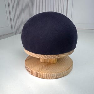 Könnte beinhalten: Eine dunkel marineblaue, stoffbezogene Kuppelform auf einem runden, hellen Holzsockel. Die Kuppel hat eine glatte, gleichmäßige Textiloberfläche, während der Holzständer eine sichtbare Maserung und ein stabiles, flaches, rundes Fundament aufweist.