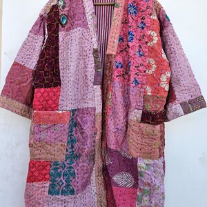 May include: A patchwork kimono-style robe in shades of pink, purple, and burgundy. The robe features a variety of patterned fabrics, including floral and geometric designs. It has long sleeves and a striped interior lining. A decorative pin is attached to the front.