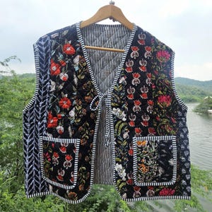 Puede incluir: Un chaleco acolchado con un estampado floral en rojo, blanco y verde sobre un fondo negro. El chaleco tiene un interior a rayas y un panel lateral con estampado en blanco y negro contrastante. Cuenta con dos bolsillos delanteros y un cierre de lazo.