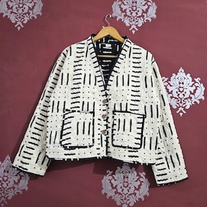 Könnte beinhalten: Cremefarbene Steppjacke mit schwarzem geometrischen Muster. Die Jacke hat einen V-Ausschnitt, zwei Taschen und Holzknöpfe. Das Futter ist schwarz, und die Jacke hängt an einem Holzbügel.