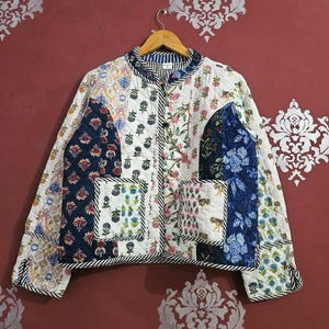 Puede incluir: Chaqueta acolchada con diseño patchwork. Presenta una mezcla de estampados florales y geométricos en blanco, azul marino y otros colores. Tiene cuello y ribetes a rayas. Colgada en una percha de madera.