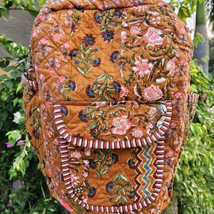 Op de afbeelding: Een gewatteerde rugzak in een warme bruine tint, versierd met een bloemenpatroon in roze, blauw en groen. De rugzak heeft twee zijzakken en een voorzak met een zigzagontwerp. De bandjes en randen zijn afgewerkt met een streepjespatroon.