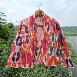 Puede incluir: Chaqueta acolchada colorida con un vibrante estampado abstracto en tonos naranja, morado, rosa y verde azulado. La chaqueta tiene cuello mandarín y está abotonada en la parte delantera. Cuelga de una percha de madera.