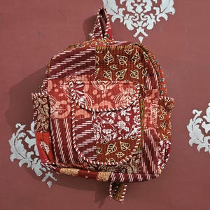 May include: A patchwork backpack in shades of red, burgundy, and cream. The bag features a variety of patterned fabrics, including leaf and floral designs. White and red piping outlines the flap and pockets. The bag has a top handle and side pockets.