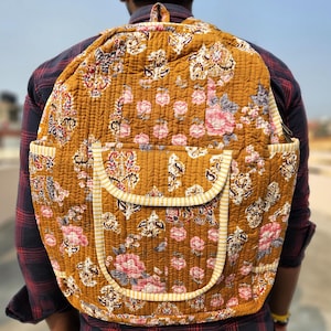 Peut inclure: Sac à dos matelassé de couleur moutarde avec un motif floral rose, blanc et bleu. Le sac à dos a une poche avant avec une bordure rayée et deux poches latérales. Le sac est porté sur le dos.
