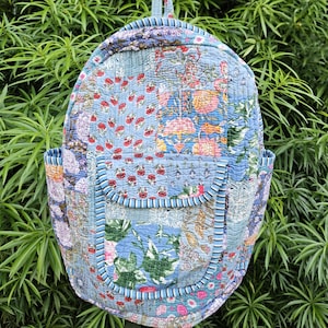 Op de afbeelding: Gewatteerde rugzak met een patchwork-ontwerp. De rugzak heeft verschillende bloempatronen in blauw, roze en rood. Het heeft twee zijzakken en een voorvak met een gestreepte rand. De rugzak heeft een handvat aan de bovenkant.