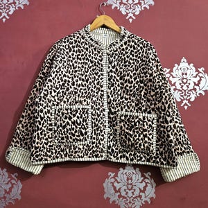 Può includere: Giacca con stampa leopardata con collo tondo e due tasche frontali. La giacca ha uno sfondo color crema con macchie nere. I bordi e le finiture sono accentuati con un tessuto a righe crema e nero.