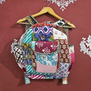 Op de afbeelding: Een patchwork rugzak met een verscheidenheid aan kleurrijke stofpatronen, waaronder bloemen- en geometrische ontwerpen. De rugzak heeft een voorvak en hangt aan een houten hanger. Het kleurenpalet omvat rood, blauw, groen en roze.