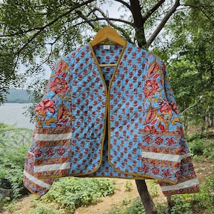 May include: A light blue quilted jacket with a floral pattern in red and gold. The jacket has a V-neck, long sleeves, and a gold trim. The jacket is hanging on a wooden hanger.