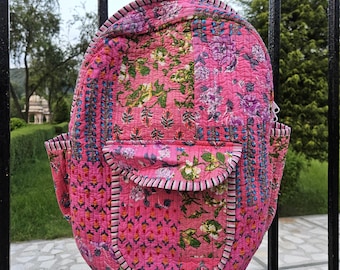 May include: A pink quilted backpack with a floral pattern. The backpack has a front pocket, side pockets, and a top handle. The floral design includes pink, purple, green, and blue hues. The backpack is hanging on a black metal fence.