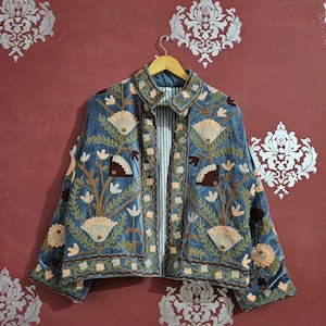 Peut inclure: Veste en velours bleu avec broderies florales complexes dans les tons pêche, marron et vert. La veste a un col et est suspendue à un cintre en bois. Le fond est un mur bordeaux avec des motifs décoratifs blancs.