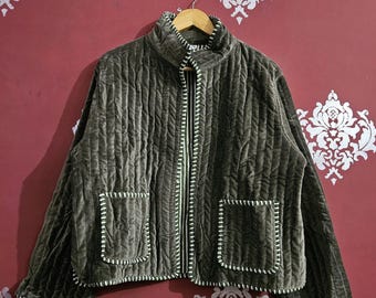 以下が含まれることがあります： ハイカラーと2つのフロントポケットが付いた、濃いオリーブグリーンのキルティングジャケット。 ジャケットは垂直のリブ模様で、白いステッチでトリミングされています。 白い花の模様が入った赤い壁に、木製のハンガーに掛けられています。