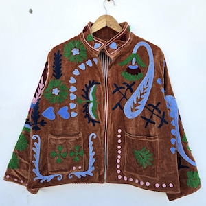 May include: Brown velvet jacket with colourful embroidered floral and paisley designs. The jacket has a collared neckline, two front pockets, and a full-length zip. The embroidery includes shades of blue, green, pink, and white.
