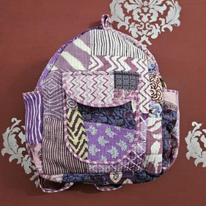 Peut inclure: Sac à dos patchwork aux tons violets, lavande et blancs. Le sac présente une poche à rabat et des poches latérales, avec divers motifs de tissu, notamment des rayures, des zigzags et des imprimés floraux. Le sac a des bretelles réglables.