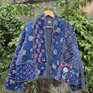 Può includere: Giacca blu trapuntata con stampa floreale e tigrata. La giacca presenta una fodera interna a righe ed è appesa a una gruccia di legno. Il design patchwork presenta accenti rossi, bianchi e blu.