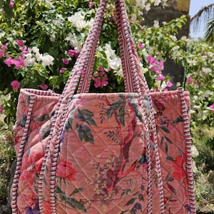 May include: A quilted tote bag in a dusty rose pink hue featuring a floral pattern with green leaves and dark accents. The bag has two shoulder straps and edges finished with a black and white striped trim, creating a structured appearance.