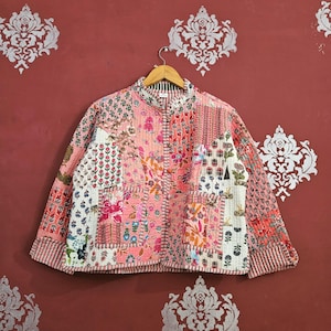 Peut inclure: Veste patchwork aux tons rose, blanc et rouge, avec des motifs floraux et géométriques. La veste a un col mandarin, des manches longues et une fermeture boutonnée. La veste est suspendue à un cintre en bois.