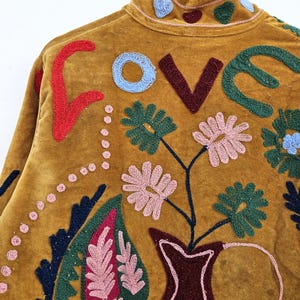 以下が含まれることがあります： マスタード色のジャケット、刺繍入り。 背中には「LOVE」の文字が赤、青、茶色、緑で刺繍されています。 ピンク、緑、青の花柄と葉柄がジャケットを飾り、ボヘミアンな美しさを演出しています。
