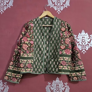 Puede incluir: Chaqueta acolchada con estampado floral en tonos verdes, rosas y crema. La chaqueta tiene cuello en V y está colgada de una percha de madera. El diseño floral se repite en toda la chaqueta.