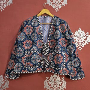 May include: Navy blue quilted jacket featuring a floral design in red and white. The jacket has two front pockets and a striped lining. It is fastened at the front with a black and white striped ribbon.