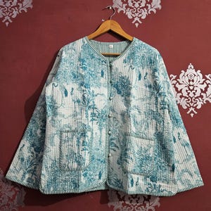 Könnte beinhalten: Gesteppte Jacke mit botanischem Druck in Weiß- und Türkistönen. Die Jacke hat einen runden Kragen, einen Knopfverschluss, zwei Vordertaschen und grüne Streifen. Die Jacke hängt an einem Holzbügel.